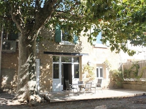 A rustic stone building with a covered patio, outdoor seating, and a large tree providing shade.