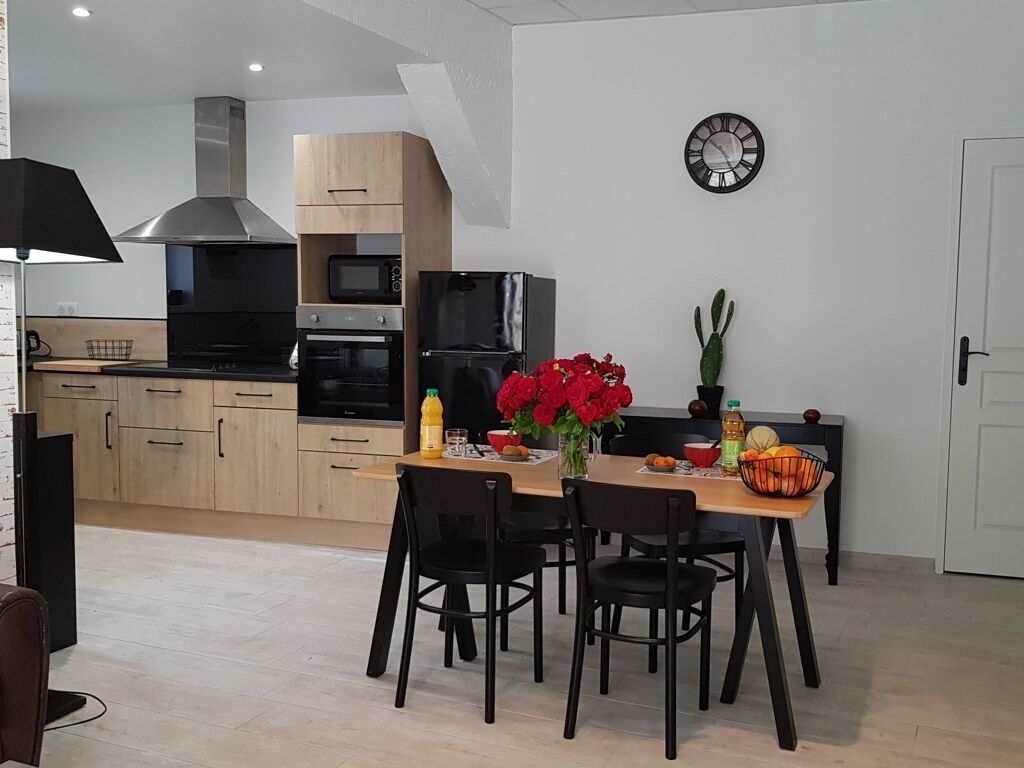 Een moderne keuken met houten kastjes, een roestvrijstalen afzuigkap en een inbouwoven. Er staat een eettafel met stoelen, een vaas met rode bloemen en een klok aan de muur.