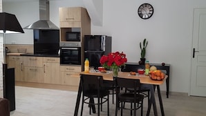 Cozinha moderna com armários de madeira, exaustor de aço inoxidável e forno embutido. Há uma mesa de jantar com cadeiras, um vaso de flores vermelhas e um relógio na parede.