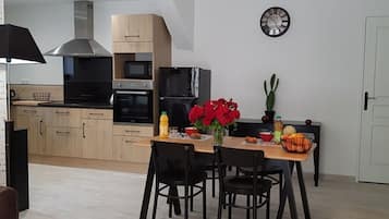 Een moderne keuken met houten kastjes, een roestvrijstalen afzuigkap en een inbouwoven. Er staat een eettafel met stoelen, een vaas met rode bloemen en een klok aan de muur.
