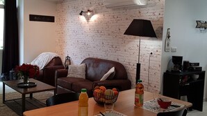 Uma sala de estar com parede de tijolos, um sofá de couro marrom, uma mesa de centro com um vaso de flores e uma mesa de jantar com uma tigela de frutas e uma garrafa de suco.