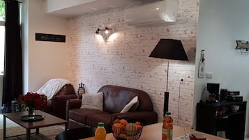 Un salon avec un mur en briques, un canapé en cuir marron, une table basse ornée d’un vase de fleurs et une table à manger avec un saladier de fruits et une bouteille de jus.