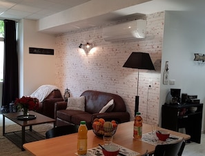 A living room with a brick wall, a brown leather sofa, a coffee table with a vase of flowers, and a dining table with a bowl of fruit and a bottle of juice.