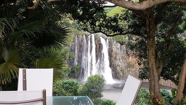 Una terraza de madera con una mesa y sillas de vidrio, con vista a una cascada y abundante vegetación.