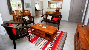 Una sala de estar con una mesa de centro de madera, dos sofás y una mesa de comedor con sillas. Se ve una cocina al fondo y una puerta a la derecha.
