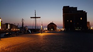 A cobblestone waterfront promenade at dusk with a historic building and a large ship docked nearby.