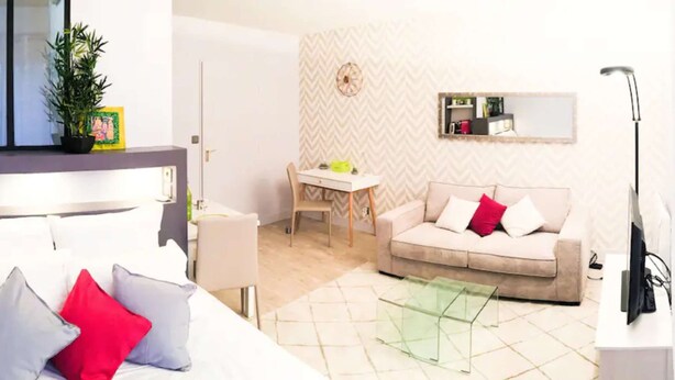 A modern living room with a white sofa, a glass coffee table, a dining table with chairs, and a small kitchen area with a mirror. - Studio Premium Toulouse Zénith - Parking,  Métro et Netflix (Toulouse)
