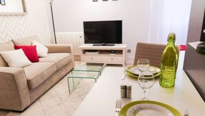 A modern living room with a sofa, a coffee table, a dining table set with plates, glasses, and cutlery. A television is placed on a stand against the wall. - Studio Premium Toulouse Zénith - Parking,  Métro et Netflix (Toulouse)