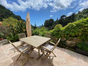 An outdoor patio with a wooden table and chairs, surrounded by lush greenery and a stone wall.