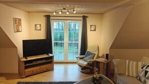A living room with a flat-screen TV, a leather armchair, a sofa, a wooden entertainment unit, and a large window with curtains.