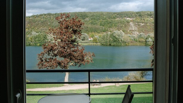 Un balcón con mesa y sillas, con vista a un lago, un árbol al frente y colinas al fondo.