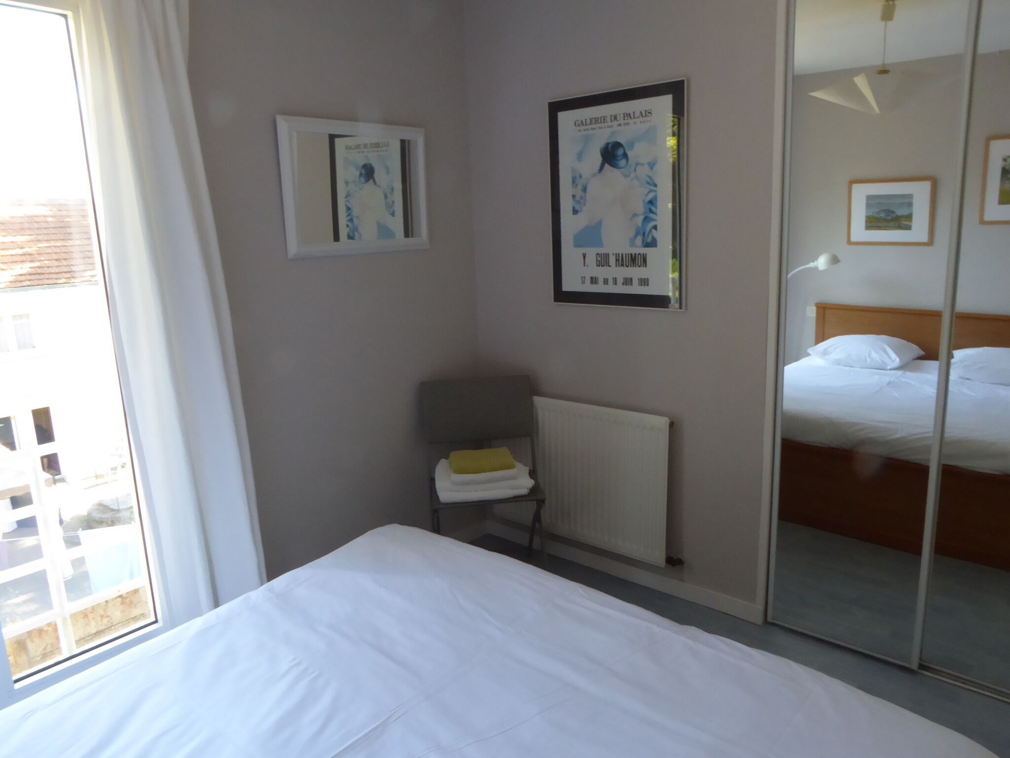 A hotel room with a bed, a radiator, a window with curtains, and two framed pictures on the wall.