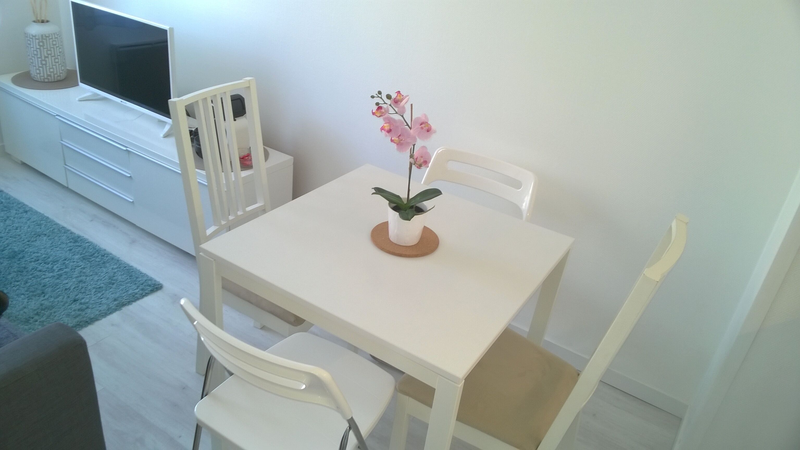 Een eethoek met een witte tafel en stoelen, een potorkidee en een televisie op een witte kast.