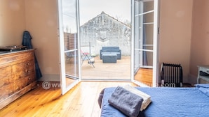 A bedroom with a wooden floor, a bed with a blue bedspread, and a view of an outdoor patio with a wooden deck and outdoor furniture. - Large atypical charming house entirely renovated. (Jonzac)