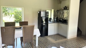 Una cocina compacta con un refrigerador negro, un microondas y una cafetera. Hay una zona de comedor con sillas de mimbre y una mesa blanca. El piso tiene baldosas estampadas y el techo es inclinado con vigas a la vista.