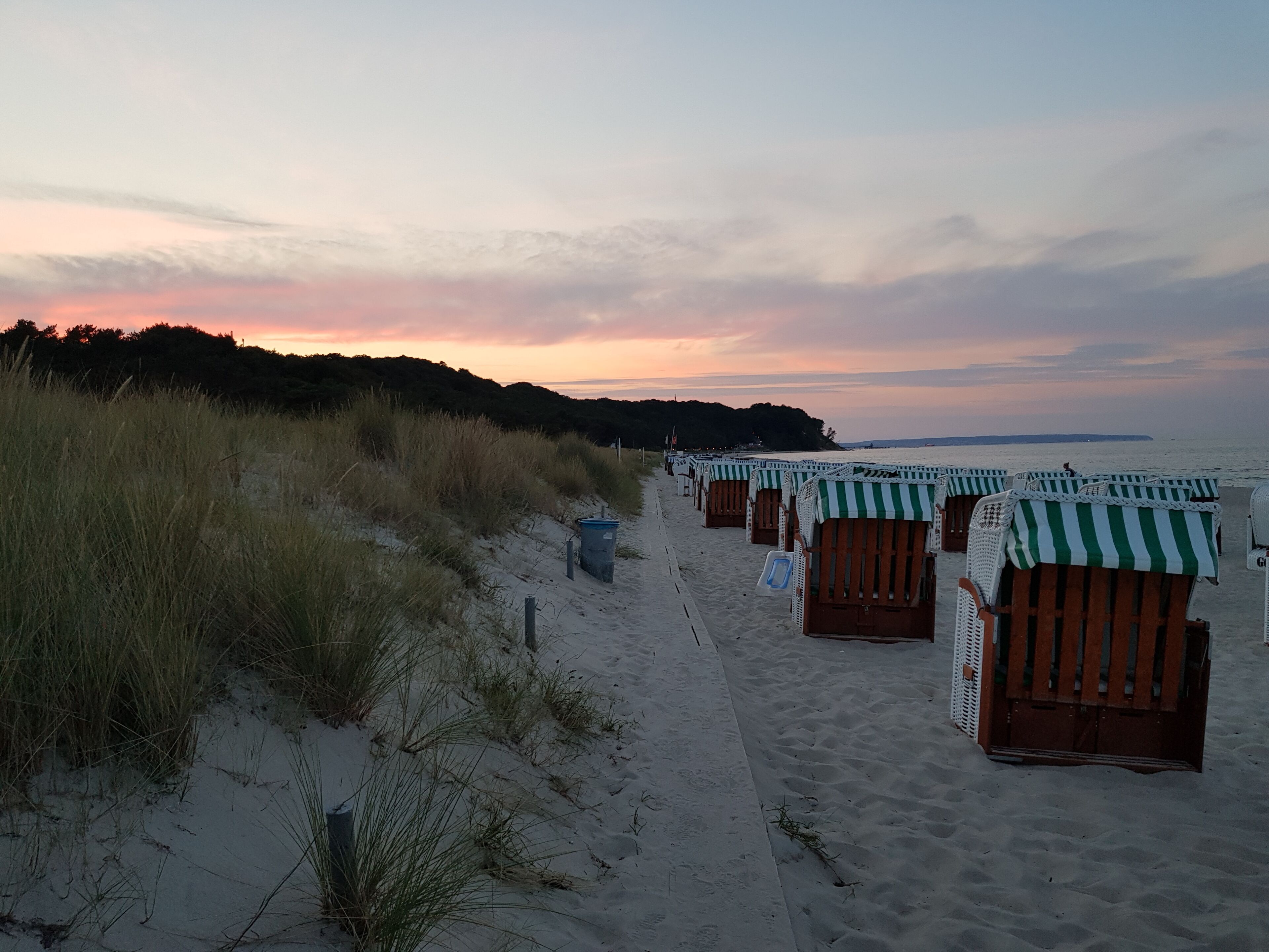 Ein Strand mit Reihen von Holzliegen mit grün-weißen Streifen, einem sandigen Pfad und einem grasbewachsenen Dünenbereich.