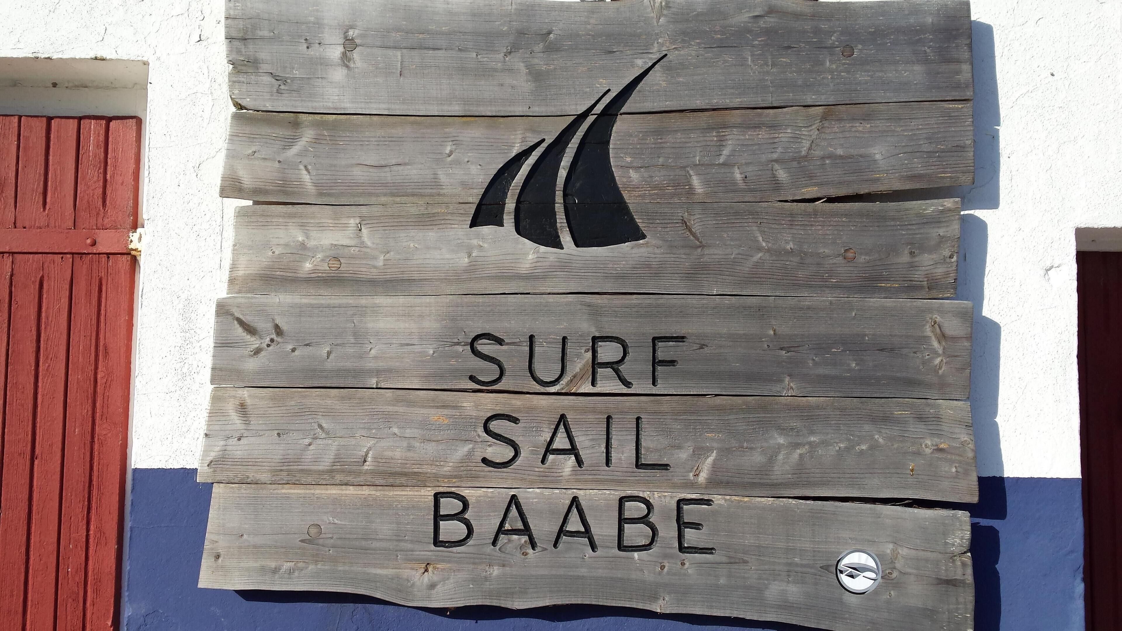 Un letrero de madera con la inscripción 'SURF SAIL BAABE' y un logotipo de velero.