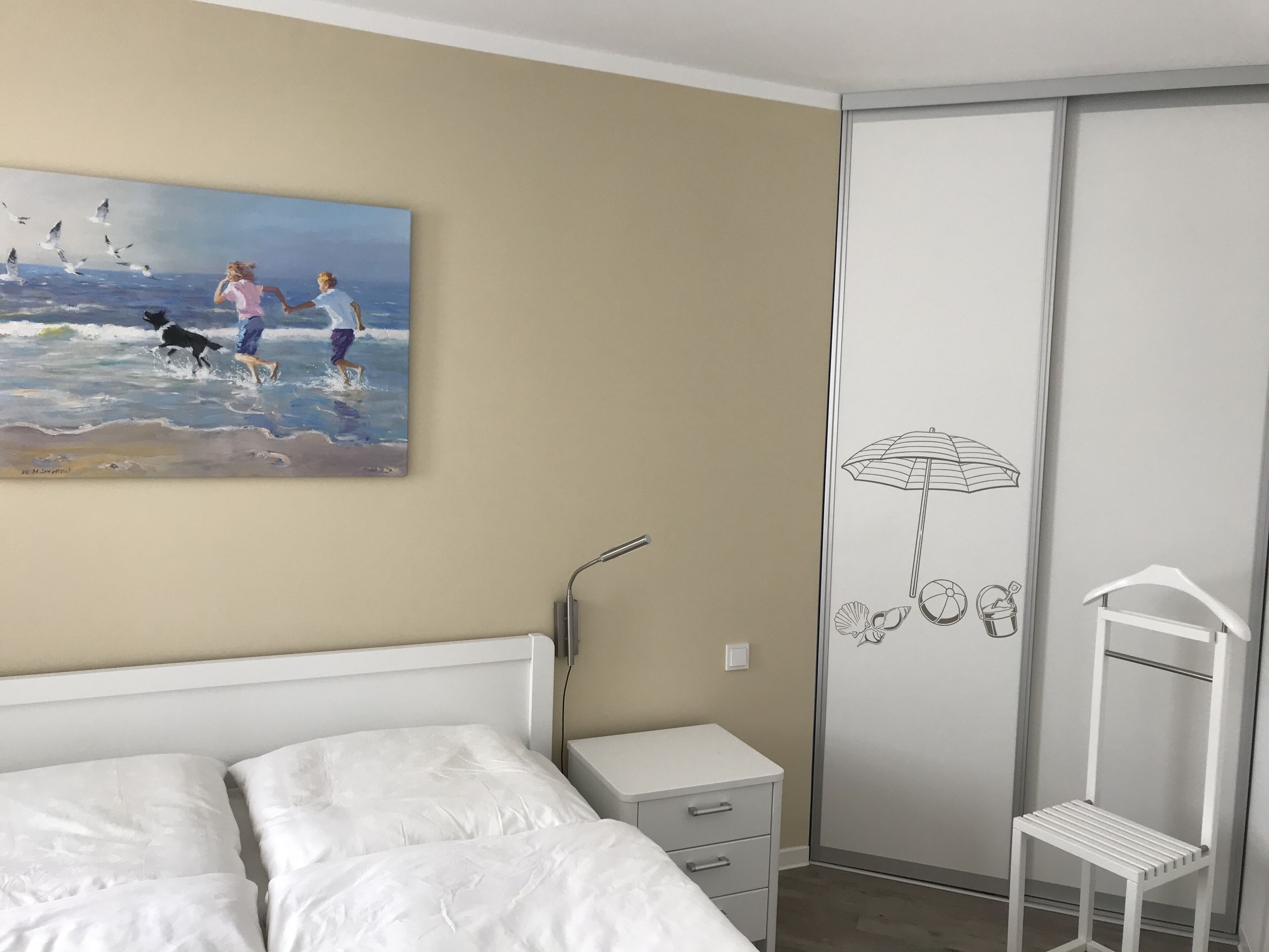 Un dormitorio con cama, mesita de noche, lámpara de pared, armario y un cuadro con motivos playeros.