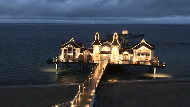 Ein Hotel am Wasser mit einer langen Pier, die sich ins Meer erstreckt, mit mehreren Gebäuden mit Rundbogenfenstern und einer zentralen Kuppelstruktur.