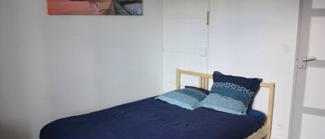 A single bed with a wooden frame and a dark blue bedspread, a white door to the right, and a painting of a coastal scene on the wall.