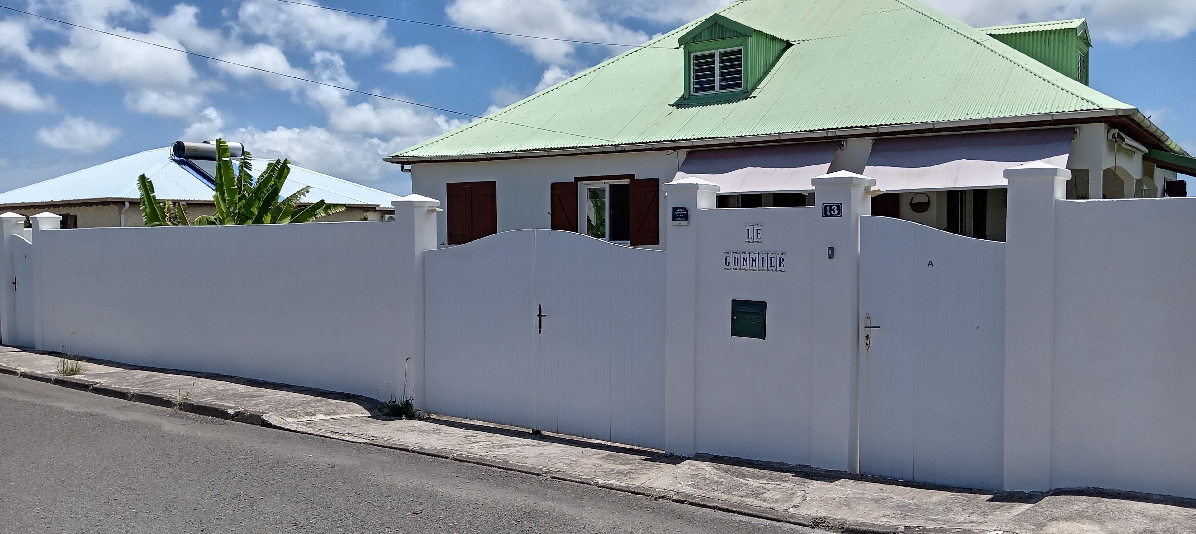 A white building with a green roof, a gated entrance, and a sign reading 'LE GOMMIE'.