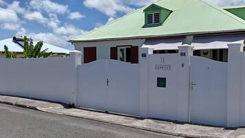 A white building with a green roof, a gated entrance, and a sign reading 'LE GOMMIE'.