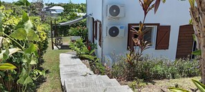 A pathway leading to a white building with two air conditioning units, surrounded by lush greenery and a small table. - Villa Le Gommier - Rental in Le Moule, Guadeloupe (LE MOULE)