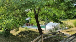 Un pont en bois mène à un espace piscine entouré d’une végétation luxuriante et à un petit bâtiment abritant une piscine couverte.