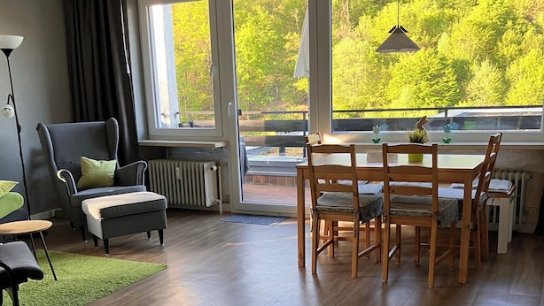 A spacious room with a wooden floor, a dining table with chairs, a grey armchair, a small table with a lamp, and large windows offering a view of greenery. - Quiet vacation oasis with a beautiful view over Bad Lauterberg (Bad Lauterberg)