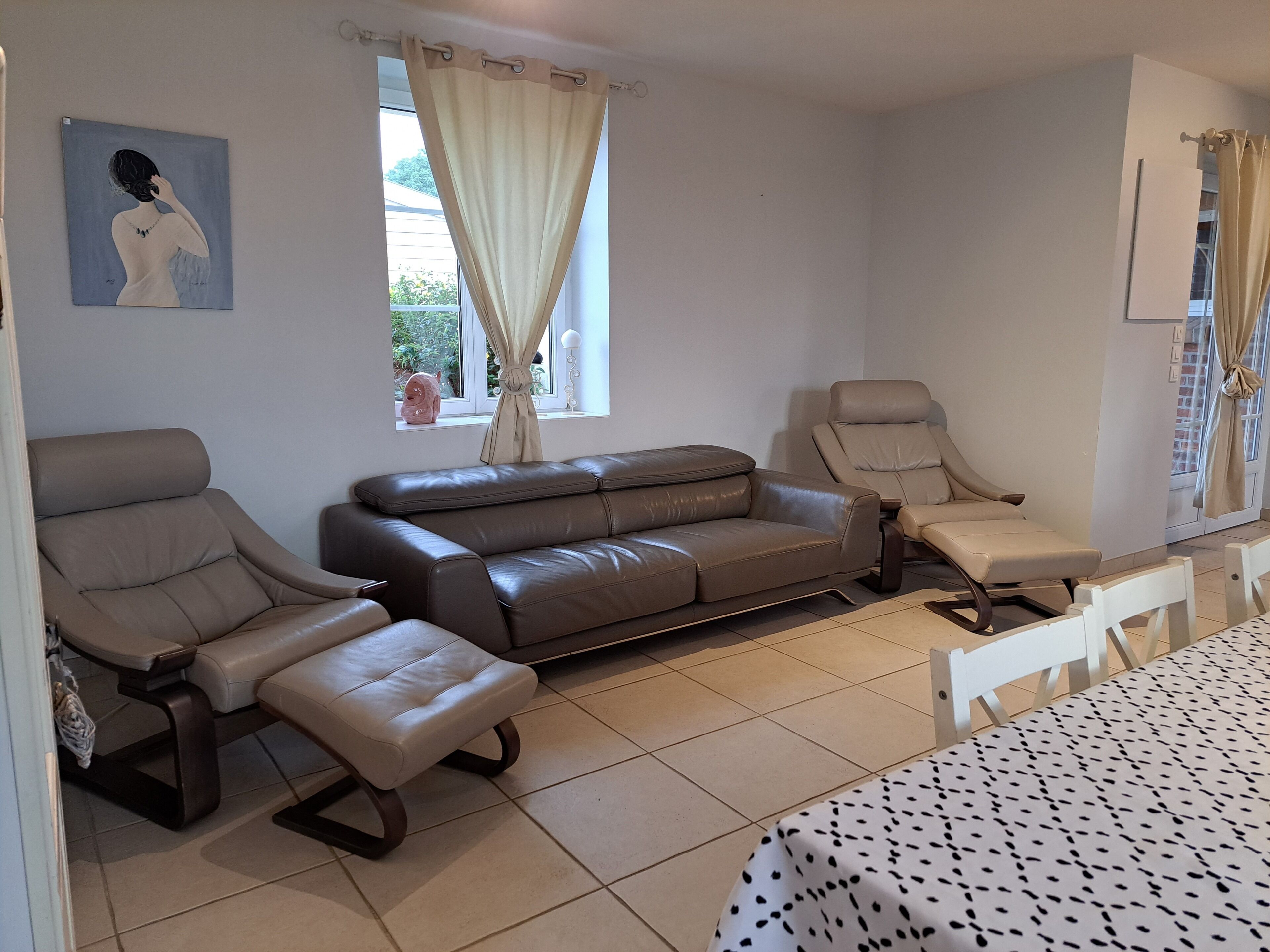 Una habitación con un sofá de cuero marrón, dos sillones color beige, una mesita blanca pequeña y una colcha estampada.