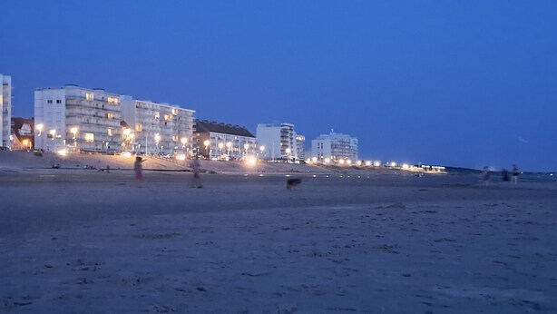 Un hotel frente a la playa con varios pisos y balcones, una playa arenosa en primer plano y un cielo despejado.
