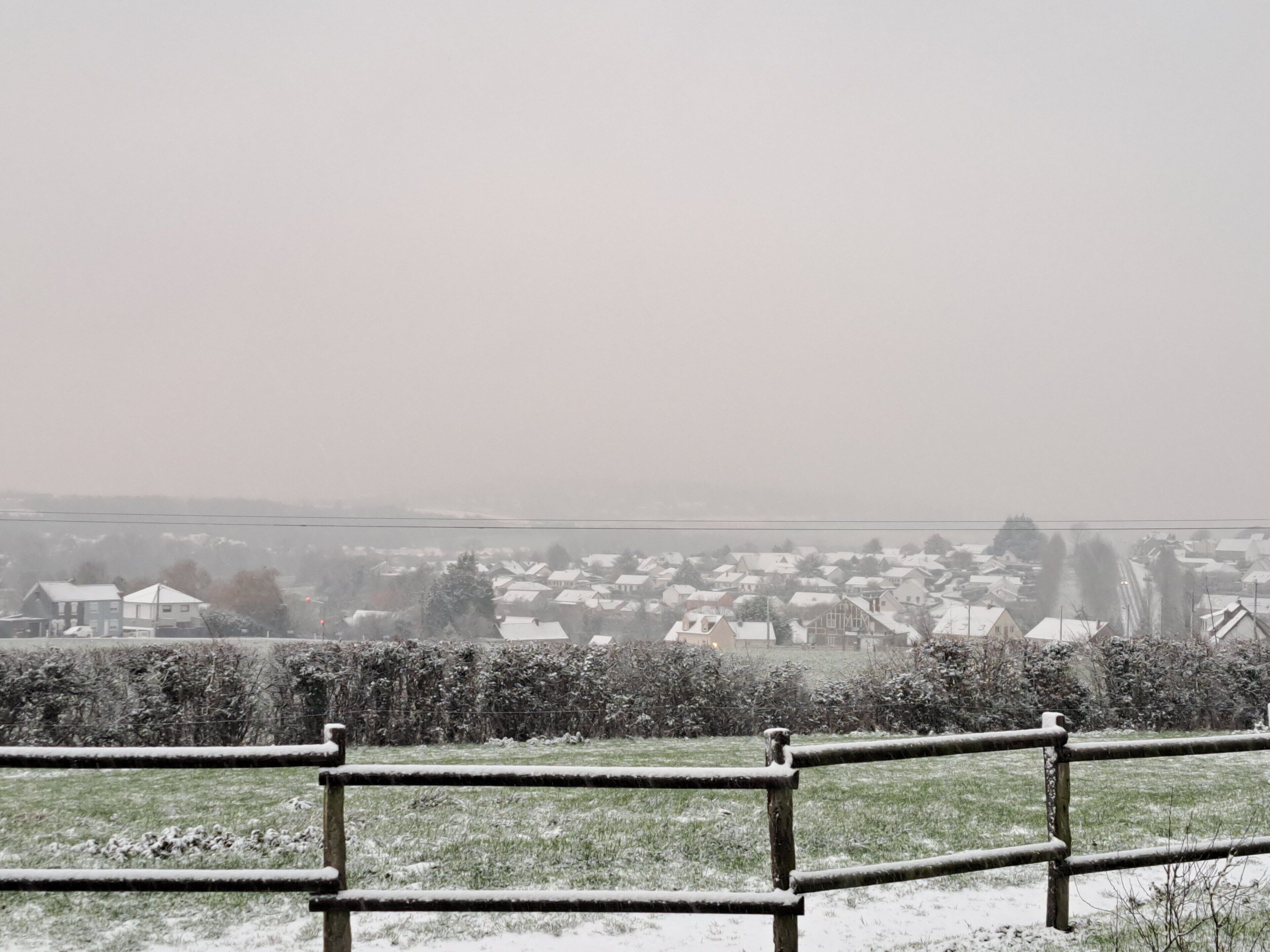 A snowy landscape with a fence in the foreground and houses in the background.