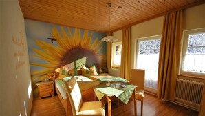 A cozy room with a wooden ceiling, a large sun mural on the wall, a bed with a green bedspread, a small table with a green tablecloth, and a window with curtains.