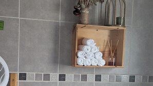 A bathroom with grey tiles, a wooden shelf with rolled towels, a fan, and a vase with dried flowers.