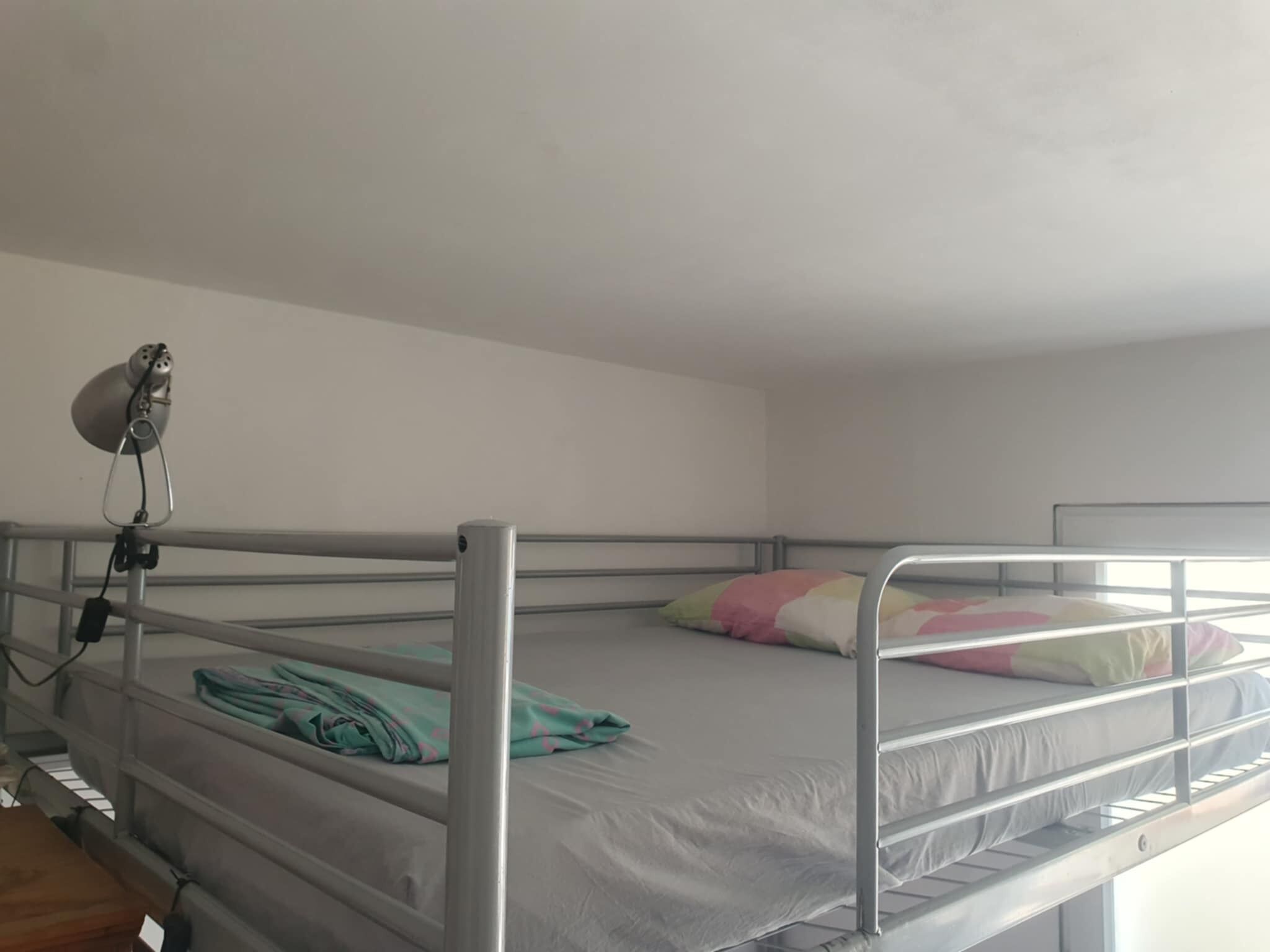Habitación con una litera de metal, un escritorio pequeño con una lámpara y una ventana que deja entrar luz natural.