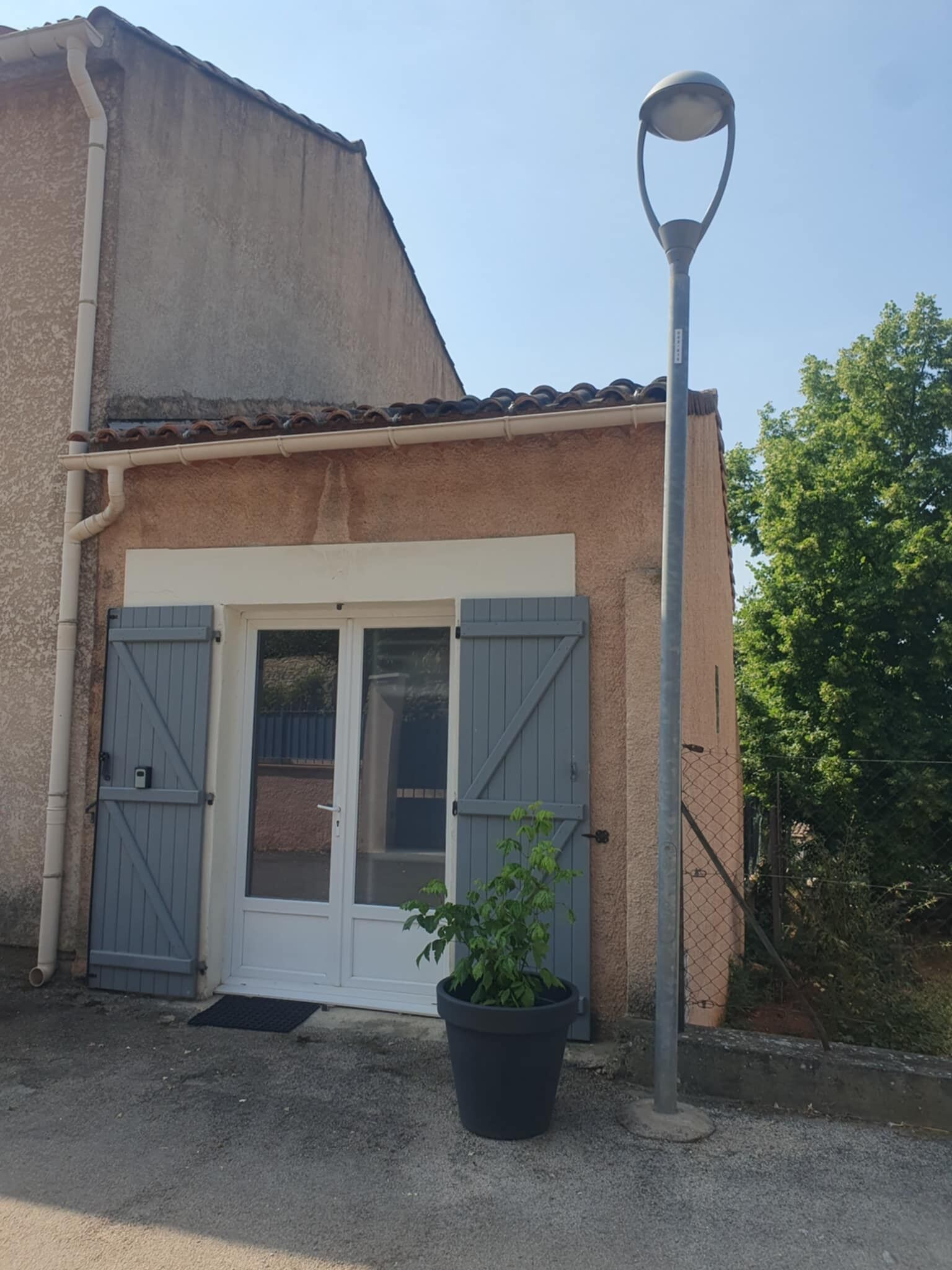 Rakennuksessa on valkoinen ovi ja siniset ikkunaluukut, sen edessä on ruukussa oleva kasvi ja oikealla puolella katulamppu.