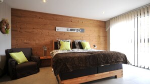 A modern bedroom with a wooden headboard, a large bed with patterned bedding, a side chair, and a bedside table with a lamp. - Cottage "Esprit Cozy" 2/4 people (Ostheim)