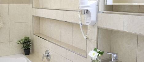Une salle de bain moderne avec une baignoire, un lavabo et un miroir. On y trouve un sèche-cheveux et une petite plante en pot, accrochée au mur.