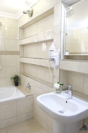 Une salle de bain moderne avec une baignoire, un lavabo et un miroir. On y trouve un sèche-cheveux et une petite plante en pot, accrochée au mur.