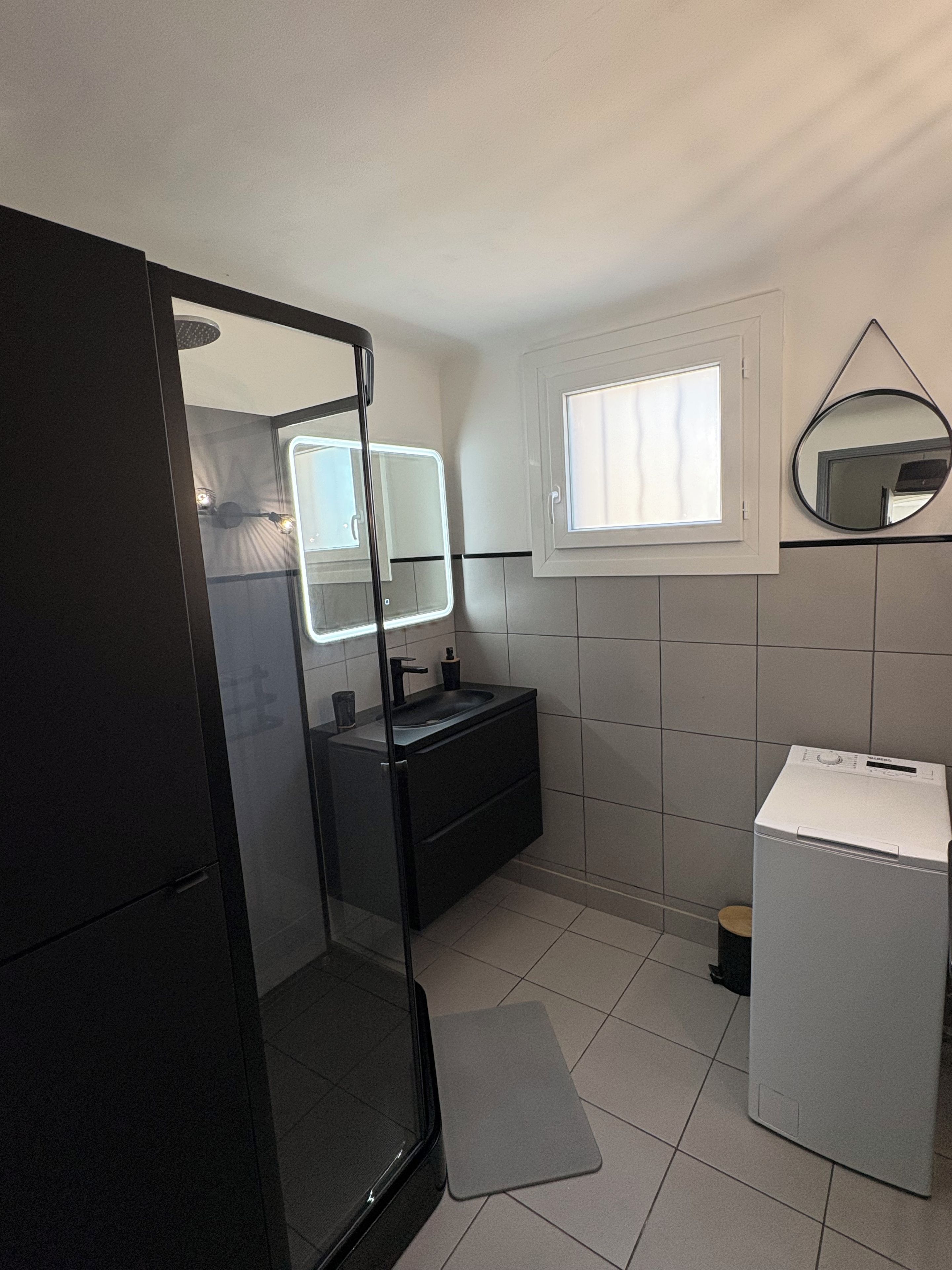 A bathroom with a glass-enclosed shower, a sink with a mirror, a window, and a washing machine.