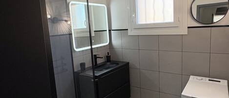 A bathroom with a glass-enclosed shower, a sink with a mirror, a window, and a washing machine.