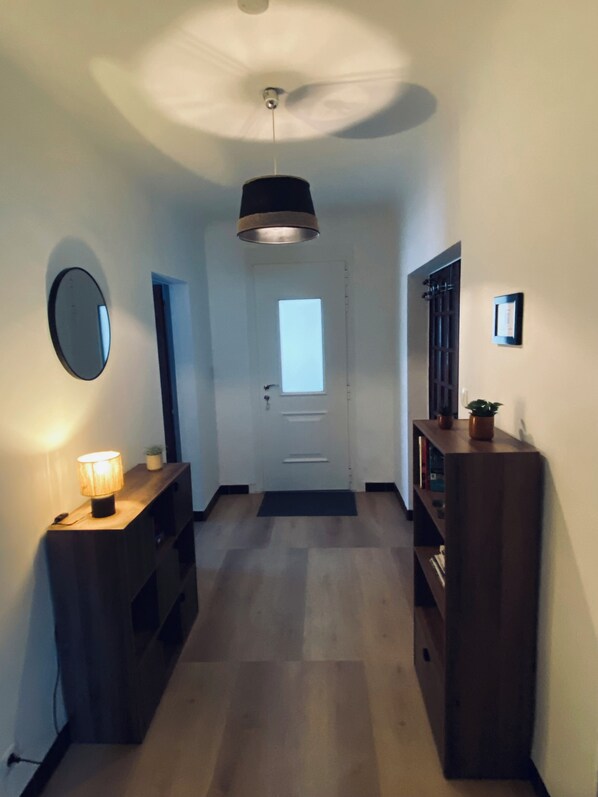 Un pasillo con ventilador de techo, una puerta con ventana, un espejo montado en la pared, una consola de madera con una lámpara y una estantería.