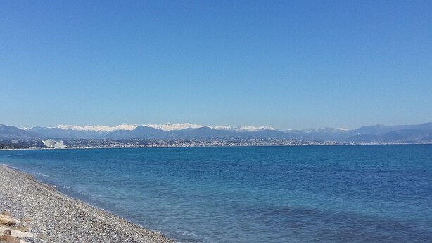 Une vue sur la côte, avec une plage de galets, une mer calme et des montagnes enneigées à l’horizon.