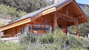 Une cabane en bois, dotée d’un toit à pente, entourée d’une végétation naturelle et avec des montagnes en arrière-plan.