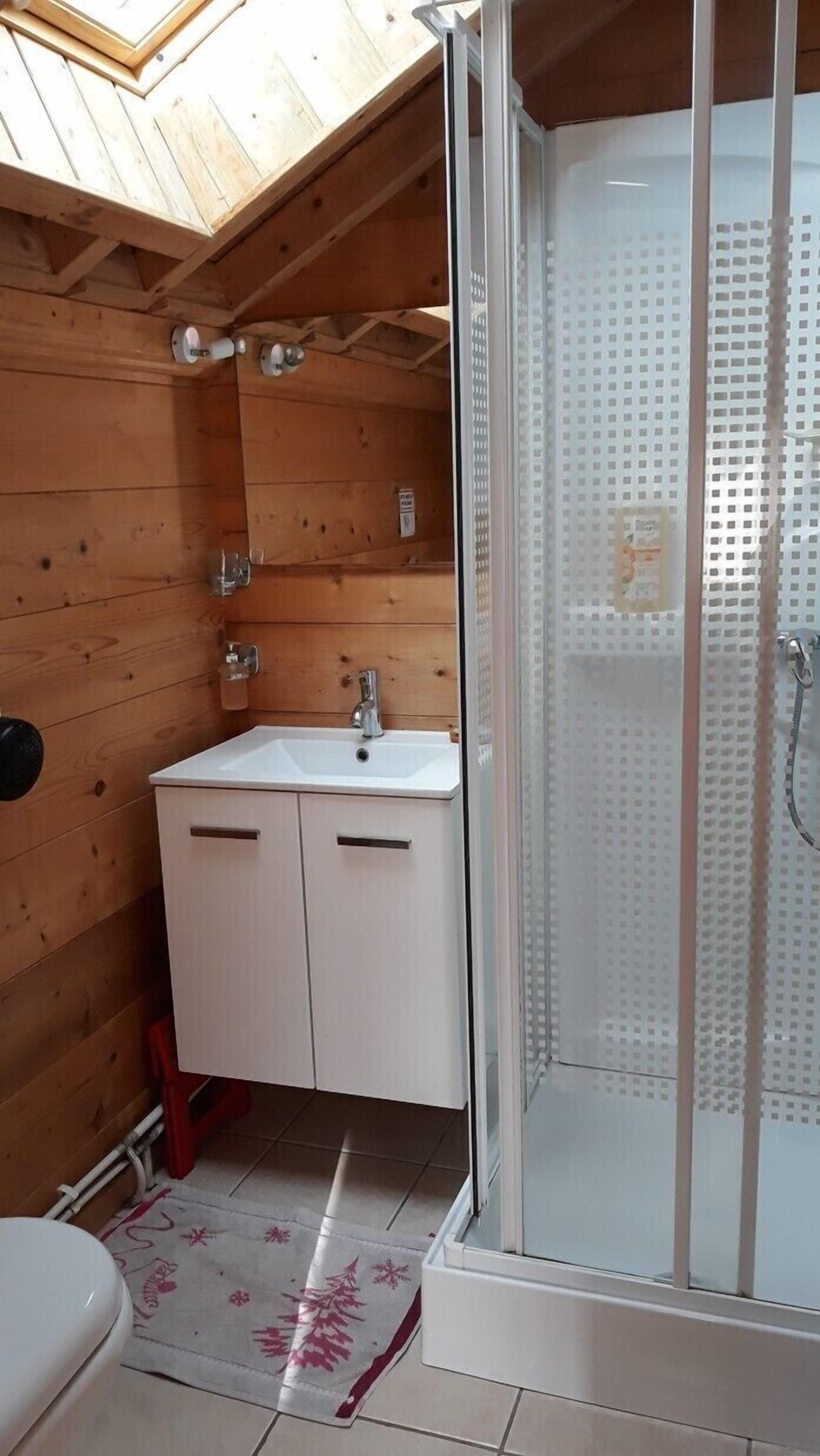A small, wooden cabin bathroom with a white sink, two cabinets, a toilet, and a glass-enclosed shower.