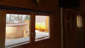 A wooden cabin with two windows, a wooden deck, and a hot tub visible through the windows.