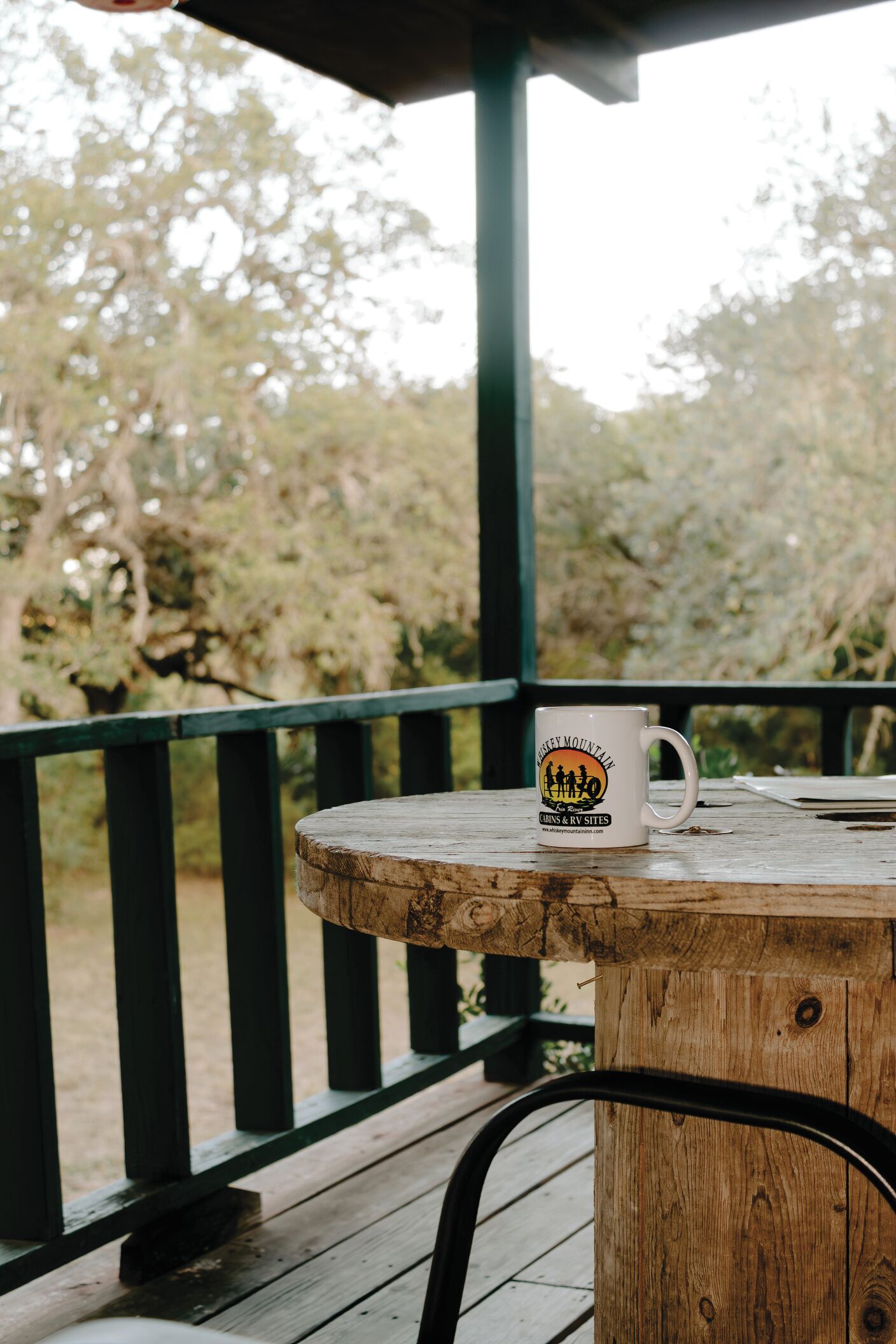 A wooden table with a mug on it, a metal chair, and a view of trees through a railing.