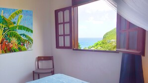 Ein Schlafzimmer mit einem Bett, einem Sessel, einem Bild und einem Fenster mit Blick auf Meer und Hügel.
