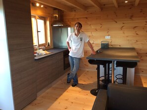A modern kitchen with wooden walls, a built-in stove, and a dining table with a black tablecloth. - Wooden chalet with sea view (Roscanvel)