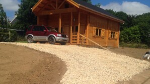 Une cabane en bois avec une allée en gravier et un SUV rouge garé devant.